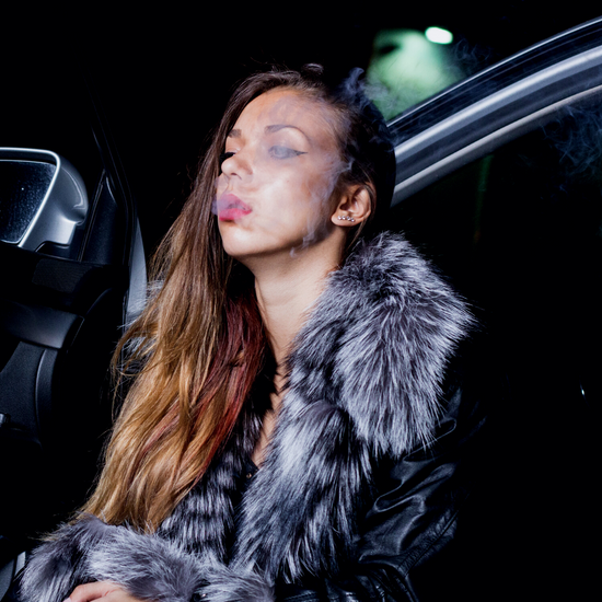 Smoke coming out of women's mouth while sitting in the drivers seat, legs on pavement with the car door open.
